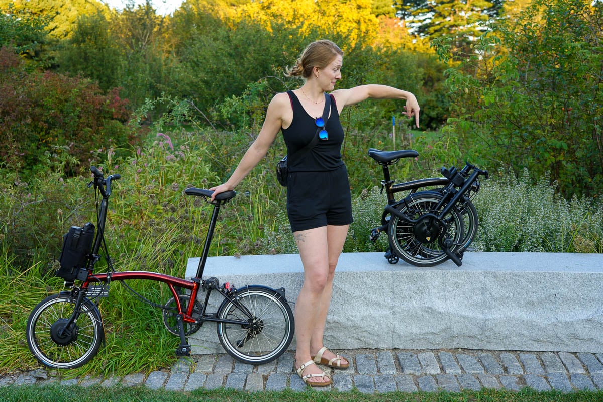 The review author, Ketti Wilhelm, stands between two collapsible Brompton electric C-Line bikes, one is folded up for storage, the other is unfolded and ready to ride. ©KettiWilhelm2025