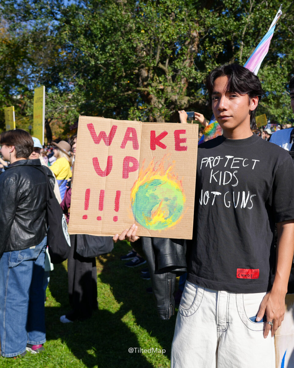 A young man holds a sign reading "wake up!!!" with a drawing of the Earth on fire, at the No Kings protest against President Donald Trump, ICE and climate change in Boston in October, 2025. ©KettiWilhelm2025
