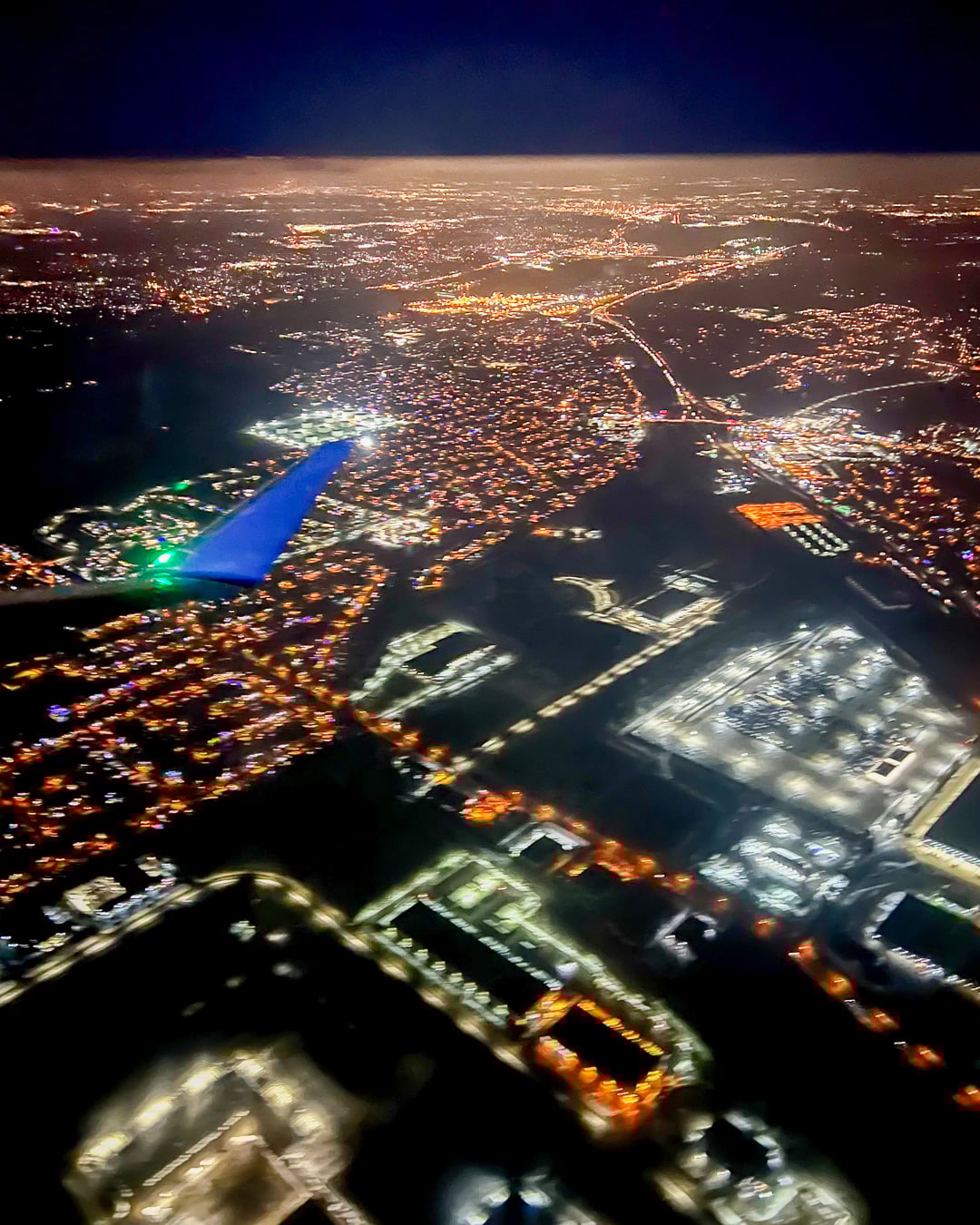 Window seat view looking out the window of an airplane at night flying over a city with orange and white city lights and a dark blue sky at the horizon. ©KettiWilhelm2026