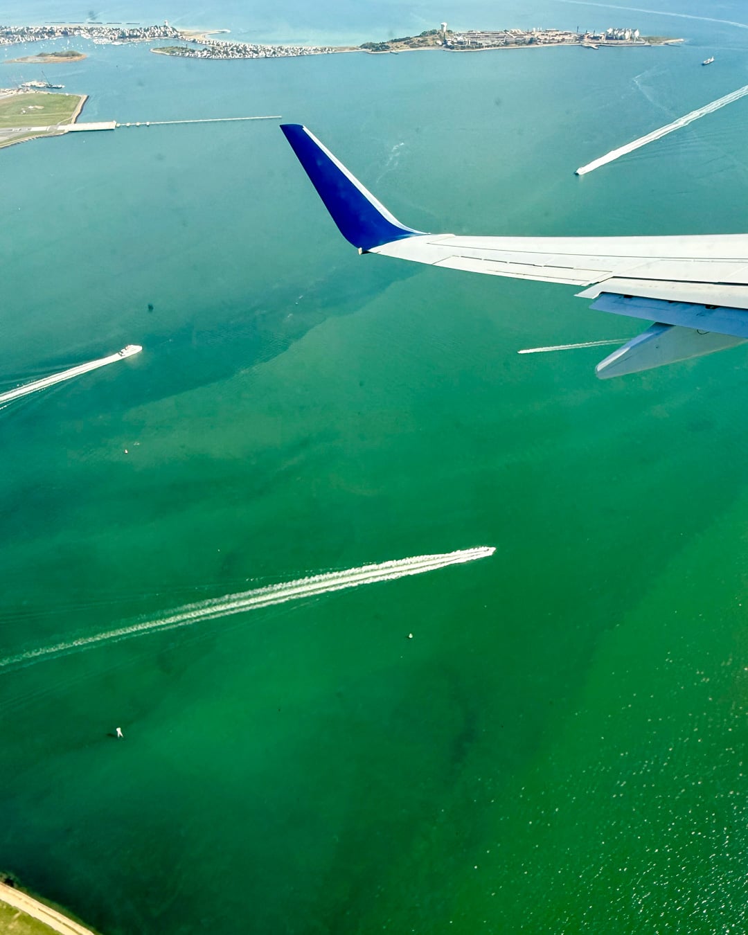 Looking down from an airplane window seat at boat wakes that look similar to jet airplane contrails in the green waters of Boston Harbor. ©KettiWilhelm2026