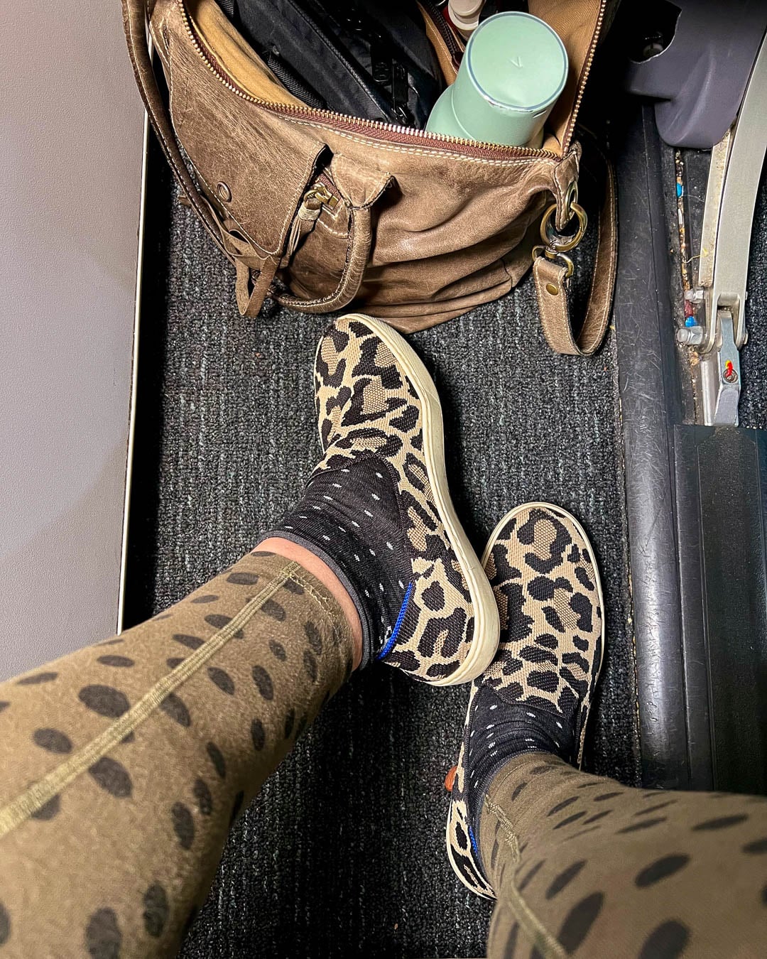The sustainable travel writer Ketti Wilhelm's feet on an airplane wearing leopard print slip-on shoes, dotted black socks, and green leggings with black dots, with a reusable water bottle that purifies tap water with UV light, removing the need to buy plastic bottles of water. ©KettiWilhelm2026