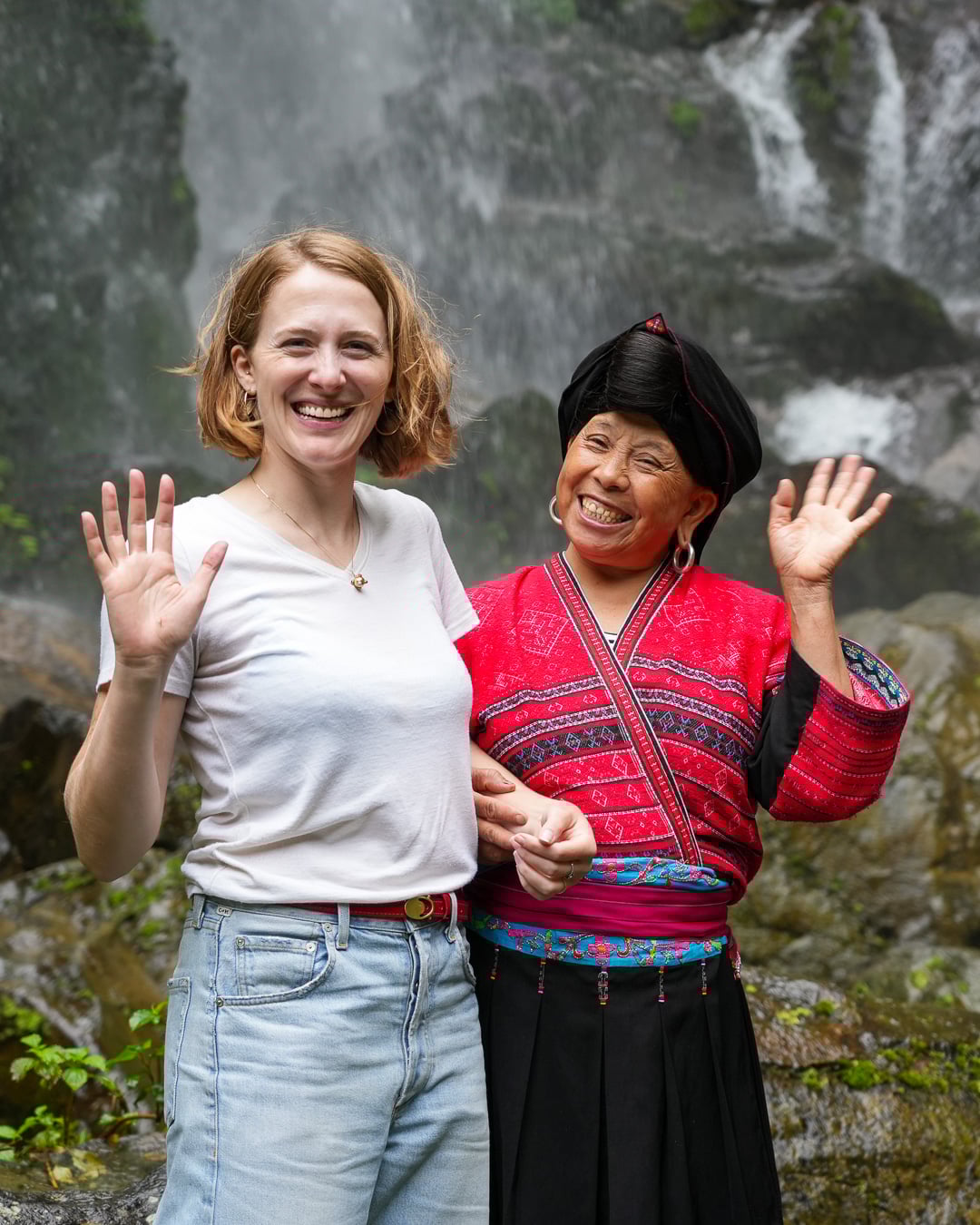 Sustainable travel writer and author of the sustainable travel blog Ketti Wilhelm stands with an indigenous Red Yao woman smiling and waving in front of a waterfall, during a tour of Longsheng, a rural China travel destination. ©KettiWilhelm2025