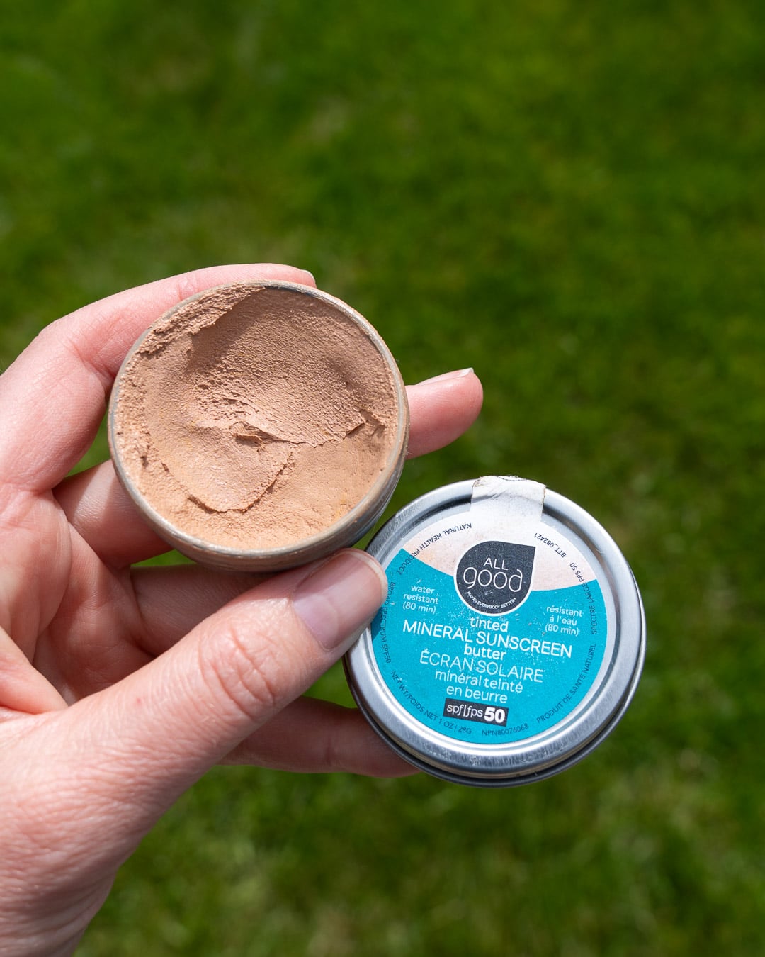 Holding a metal tin of All Good's tinted mineral SPF 50 sunscreen in my hand in front of a green lawn. ©KettiWilhelm2026
