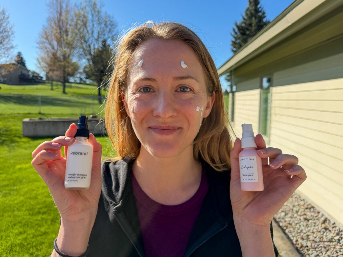 The review writer and sunscreen tester Ketti Wilhelm, a white woman i her 30s, holding bottles of mineral face sunscreen from Earth Harbor and Rebrand Skincare, with blobs of both of her face, getting ready to compare the two on opposite of her face. ©KettiWilhelm2026