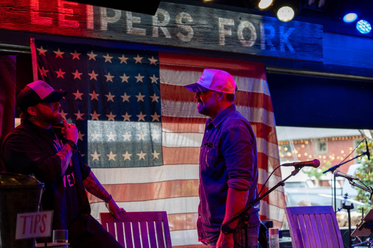 Two white men wearing baseball caps, including famous Nashville-based country rock singer Brantley Gilbert, chat on stage at the Fox and Locke Restaurant, famous for its open mic music show on Thursday nights, in Leiper's Fork, Tennessee, in front of a large US flag. ©KettiWilhelm2026