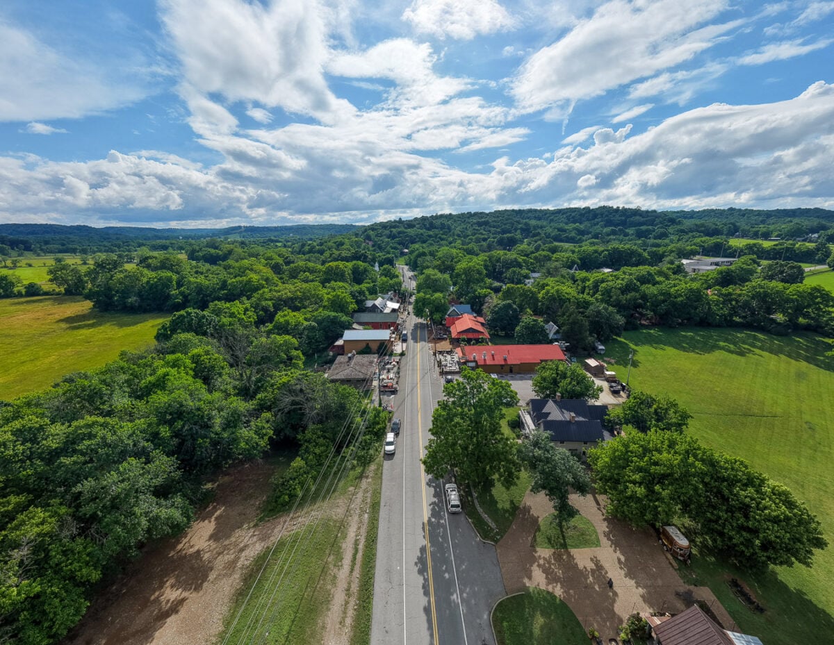 Aerial view of the village of Leiper's Fork, Tennessee, with a single road though the town and a few red-roofed buildings, surrounded by green trees, farm fields, and fluffy white clouds in the background. ©KettiWilhelm2026