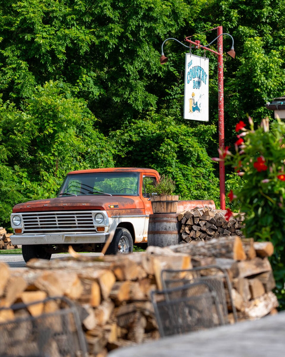 A vintage truck parked outside the Country Boy Restaurant in Leiper's Fork, Tennessee, with green trees in the background and a pile of firewood in the foreground. ©KettiWilhelm2026