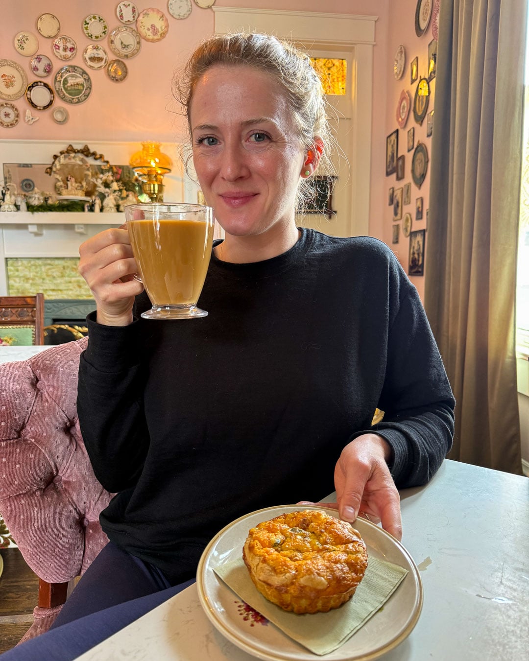 Sustainable travel writer and influencer Ketti Wilhelm, of TiltedMap.com, a white woman with blond hair wearing a black sweatshirt, holds a cup of coffee and plate with a homemade quiche at Triple Crown Bakery, one of the best breakfast spots in Franklin, Tennessee. ©KettiWilhelm2026