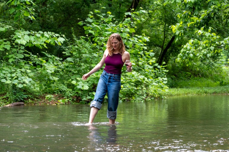 Sustainable travel writer and influencer Ketti Wilhelm, of TiltedMap.com, a white woman with blond hair walks in a stream holding a glass of red wine from local winery Crown Winery, in Leiper's Fork, Tennessee. ©KettiWilhelm2026