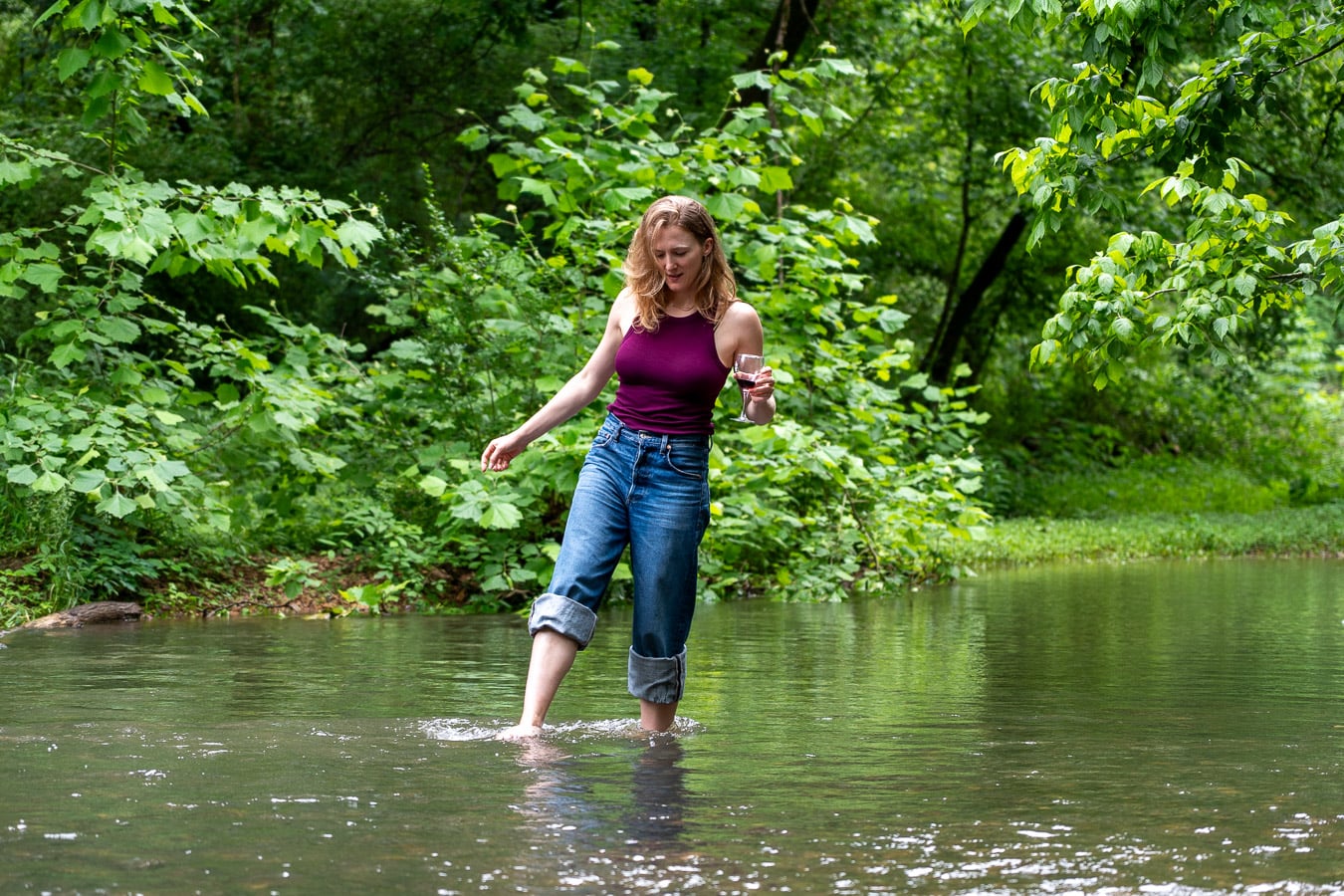 Sustainable travel writer and influencer Ketti Wilhelm, of TiltedMap.com, a white woman with blond hair walks in a stream holding a glass of red wine from local winery Crown Winery, in Leiper's Fork, Tennessee. ©KettiWilhelm2026
