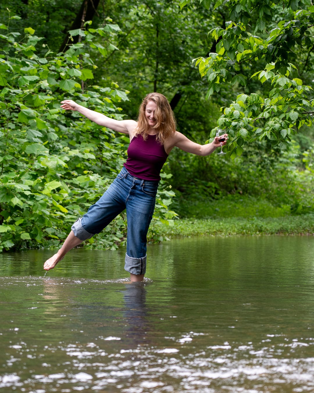 Sustainable travel writer and influencer Ketti Wilhelm, of TiltedMap.com, a white woman with blond hair walks in a stream holding a glass of red wine from local winery Crown Winery, in Leiper's Fork, Tennessee. ©KettiWilhelm2026