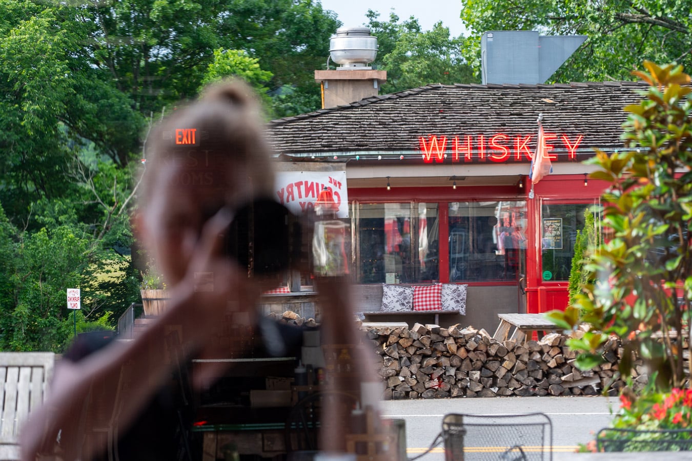 A red neon "whiskey" sign at the back of the Fox and Locke is visible in a window reflection, along with the Country Boy Restaurant across the street, and the reflection of the photographer, Ketti Wilhelm, a white woman whose face is covered by a professional camera. ©KettiWilhelm2026.